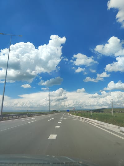 The image depicts a highway in Lusakert, Armenia, under a bright blue sky dotted with fluffy white clouds. The road is asphalt with clear white lane markings and directional arrows. On the left side of the road, a metal guardrail runs along the edge, with streetlights positioned at intervals. On the right side, a grassy field stretches out, bordered by a concrete curb and a few power poles. In the distance, a line of green hills can be seen, with a small cluster of buildings nestled at their base. A circular speed limit sign indicating "90" is visible on the right side of the road. The perspective suggests the photo was taken from a vehicle driving on the highway.