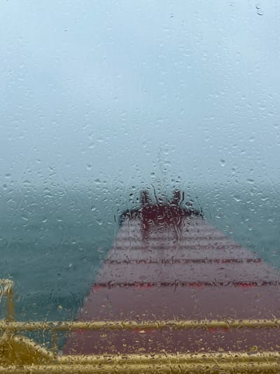 The image depicts an exterior scene observed through a window heavily streaked with numerous raindrops, indicating current precipitation. A wide expanse of dark teal water occupies the middle ground under an overcast, light blue-grey sky. In the mid-distance, a large reddish-brown vessel or structure with horizontal banding and a distinct upper section is partially visible on the water. Two dark, indistinct human figures are discernible standing on the top deck or roof of this reddish-brown object. A blurry yellow railing or barrier is present in the immediate lower foreground, suggesting the viewpoint is from another vessel or a waterfront structure. The scene is situated in the coastal waters of Bangor, United Kingdom.