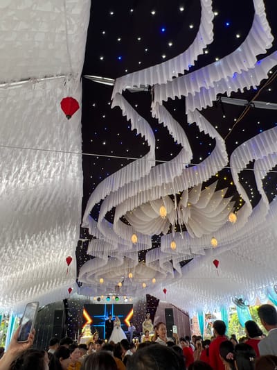 This image captures an indoor or covered event space, likely a festive celebration or reception. The ceiling is ornately decorated with flowing white fabric structures, resembling waves or stylized petals. These are interspersed with a black backdrop featuring numerous small twinkling lights in white, blue, and yellow, creating a 'starry night' ambiance. Several larger, warm-toned, lantern-like lights and small red heart-shaped or traditional lantern decorations are suspended from the ceiling.

In the foreground, a crowd of people, mostly viewed from behind, forms an audience, intently looking towards a stage. One person in the immediate foreground holds a smartphone, seemingly recording the event.

At the far end of the venue, a brightly lit stage is visible. A large LED screen on the stage displays abstract blue and orange geometric patterns. On the stage, a man in a suit and a woman in a white gown, possibly a wedding dress, are present, alongside another individual in a yellow traditional outfit. Speakers and stage lighting are also visible.

The scene suggests an evening event, possibly a wedding reception, given the attire of individuals on stage and the celebratory decorations. The event is taking place in Bình An, Vietnam. No discernible text is visible.