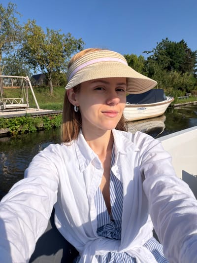 A woman is taking a selfie on a boat. She is wearing a straw sun visor with a pink band, a white button-down shirt, and a blue and white striped top underneath. She has on silver hoop earrings. The boat is on a body of water, likely a canal or river, with trees and a swing set on the shore in the background. The sky is clear and blue. The scene appears to be in a rural area, possibly Rhenoy, Netherlands, given the presence of boats and canals.