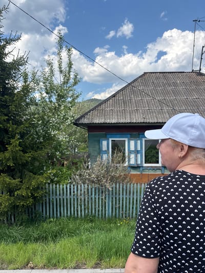 A medium shot captures a woman in a black and white patterned dress and white baseball cap standing next to a small, one-story house. The house has a gray, corrugated roof and blue window frames. A wooden fence in a matching blue color runs along the side of the house, bordered by grass. Lush green bushes and trees fill the foreground, with a sky full of white clouds visible behind the house. This location is Овсянка, Russia.
