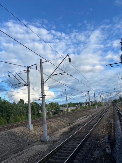 The image shows a view of railroad tracks with overhead electrical lines and poles. The tracks run parallel to each other, curving into the distance. Trees and some buildings are visible in the background. The sky is blue with scattered white clouds. The overall scene suggests an outdoor, non-urban setting, likely a rural area or a section of a railway line outside of a major city center. The presence of electrical catenary systems indicates an electrified railway.