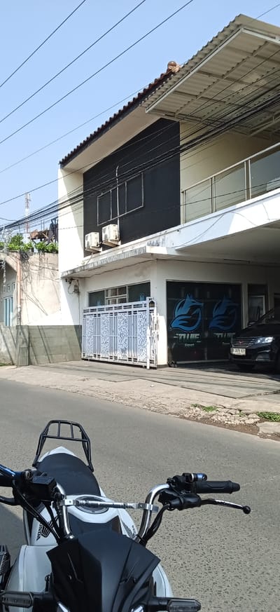 The image shows a white motorcycle parked on a street in front of a two-story building. The building has a dark colored facade on the upper level and white on the lower level. There are two air conditioning units mounted on the exterior wall of the upper level. A white metal gate is in front of the ground floor. Two blue wolf logos are visible on the windows of the ground floor. A black car is parked to the right of the gate. The street appears to be paved. The sky is clear and blue. The scene is set in the city of Ciapus, Indonesia.