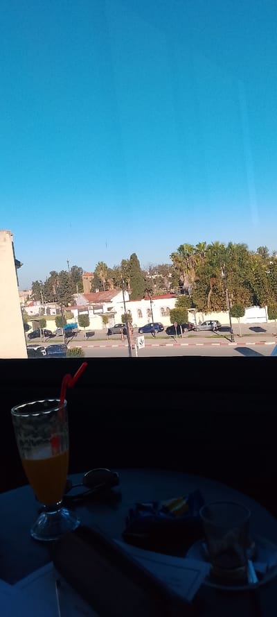 The scene depicts an outdoor urban environment in Aïn El Aouda, Morocco, viewed from an indoor setting through a window. In the immediate foreground, a dark table holds several items: a glass filled with an orange liquid and a red bent straw, a pair of dark sunglasses, a crumpled blue and yellow packet (likely snack packaging), and an empty clear glass teacup resting on a white saucer with a silver spoon. A document or menu is partially visible beneath these objects.
Beyond the window, a multi-lane asphalt road is visible, flanked by a sidewalk featuring a distinct red and white striped curb. Several vehicles are parked along the curb, including a black SUV, a light blue pickup truck, a dark blue sedan, and a silver hatchback. Street poles and lampposts are intermittently placed along the sidewalk.
In the background, a series of light-colored buildings with varying architectural details, some featuring multiple windows and gates, are visible. Dense vegetation, including numerous palm trees and other green deciduous and coniferous trees, obscures parts of the buildings. A prominent, square, light brown tower, possibly a minaret, stands out amidst the trees in the distant center-left. The sky above is clear and vibrant blue, indicating daytime conditions.