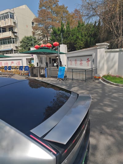 The image depicts the entrance to a facility in Jurong, China, viewed from behind a parked silver/grey car. The car's black roof and rear spoiler are prominent in the foreground. In the midground, a white wall displays red Chinese characters, "勤学实干" (Qínxué Shígàn), which translates to "Study diligently and work practically."

The entrance features a gate with metal fencing and pedestrian turnstiles. Above the entrance, three red lanterns are visible, and green canopies provide shade. Yellow and blue barricades with Chinese text are positioned in front of the entrance; one yellow barricade reads "门前区域 禁止上下客" (Area in front of the gate, no picking up or dropping off passengers), and a blue sign indicates "行人通道入口" (Pedestrian access entrance). A blue information board with white text stands near the pedestrian entrance. Several individuals are present near the turnstiles, one appearing to be a security guard in dark attire.

To the right of the entrance, a spherical bollard with yellow and red stripes marks the edge of a small landscaped area with grass and pink flowers. In the background, a multi-story, light-colored building with balconies and windows is visible, bearing the number "13" on its facade. Various trees with both green and bare branches are situated behind the entrance, under a clear blue sky. Shadows from unseen objects extend across the asphalt road in the foreground.
