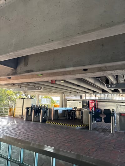 This image captures an unoccupied elevated transit station concourse in Hialeah, Florida, United States, likely a Metrorail station. The scene is primarily composed of concrete structures, a tiled floor, and automated fare collection equipment under natural lighting.

The concourse features a reddish-brown tiled floor with a square pattern. Overhead, a substantial concrete ceiling is visible, supported by large concrete beams and incorporating recessed circular light fixtures. Multiple "EXIT" signs are present: a prominent red illuminated sign with white text, and several black signs with white downward-pointing arrows indicating exits and a bus transfer point.

In the midground, a line of stainless steel automated fare gates spans the concourse. To the left, several standard turnstiles with black paddle barriers are positioned. Central to the gates is a rectangular, metallic service counter or booth, behind which a single black stool is placed. The floor area immediately in front of this counter is marked with black and yellow diagonal hazard stripes. To the right of the counter, two wider fare gates are visible, each displaying universal access symbols for wheelchairs and bicycles on their black paddle barriers. Further right, a red Coca-Cola branded vending machine is situated against a metal grated wall.

To the far left, beyond the fare gates, metal railings border the edge of the concourse, offering a view of green trees under clear daylight, indicating an open-air side. The foreground shows a low barrier with a textured, possibly translucent, surface. No individuals or specific actions are observed within the frame.