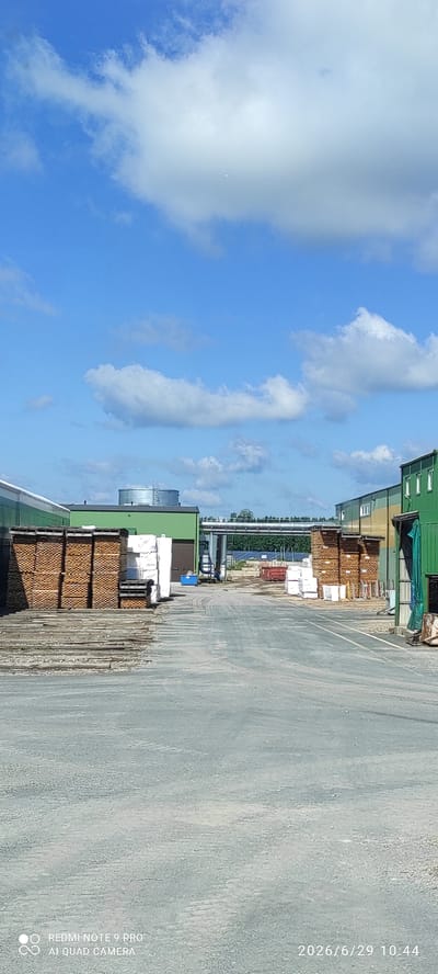 The image depicts an industrial outdoor area with several green buildings. In the foreground, there is a gravel road leading through the scene. On the left side of the road, stacks of lumber are visible, along with some bundled materials wrapped in white plastic. Further down the road, more stacks of lumber and white-wrapped items are present. On the right side, another green building with windows is visible, and a green tarp hangs near the edge. In the background, a large cylindrical tank is situated on top of a green building. Beyond the buildings, a line of trees is visible, with a bright blue sky filled with scattered white clouds overhead. The bottom right corner of the image contains a timestamp: "2026/6/29 10:44". The bottom left corner displays text indicating the device used: "REDMI NOTE 9 PRO" and "AI QUAD CAMERA". This scene is located in the city of Verijärve, Estonia.