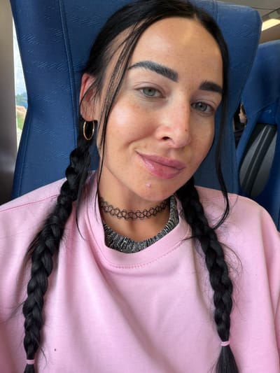 A close-up, vertical shot features a person with fair skin and dark hair styled in two braids. Their eyes are open, and they are looking towards the camera. The person has freckles across their nose and cheeks. They are wearing a light pink sweatshirt and a black choker necklace. A gold hoop earring is visible in their left ear. The background is a textured blue surface, possibly a seat or upholstery, suggesting an indoor setting like a vehicle. The lighting indicates it is daytime, with natural light entering from the left side of the frame. The image conveys a casual and relaxed mood. There is no visible text in the image.