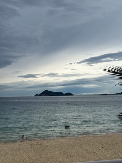 A vertical, wide shot depicts a tranquil beach scene in Song Cau, Vietnam, under a cloudy sky. The foreground features a sandy beach with scattered debris and a white railing. In the middle ground, the turquoise ocean stretches to the horizon, dotted with several small boats. Two individuals are visible wading in the shallow water near the shore. In the distance, a prominent island with a rugged, mountainous silhouette dominates the seascape, partially obscured by the overcast sky and wispy clouds. The scene conveys a sense of calm and natural beauty.