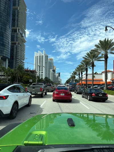This image captures a traffic jam on a sunny day in Sunny Isles Beach, Florida, USA. The perspective is from inside a bright green vehicle, looking forward through its windshield and hood, which reflects the surrounding environment. In front, a white Tesla Model Y is in the left lane, followed by a gray Chevrolet pickup truck and a red Tesla Model 3. The right lane has a black Mercedes-Benz S-Class and other vehicles further ahead.
The urban landscape is dominated by tall, modern high-rise buildings, indicative of a coastal city. Palm trees line the right side of the street, with a building featuring an orange awning visible behind them. The sky is blue with scattered white clouds. Construction cranes are visible in the background, suggesting ongoing development. The road is paved with asphalt and marked with white lane dividers. The overall scene depicts a typical urban traffic situation in a well-developed coastal city under clear skies.