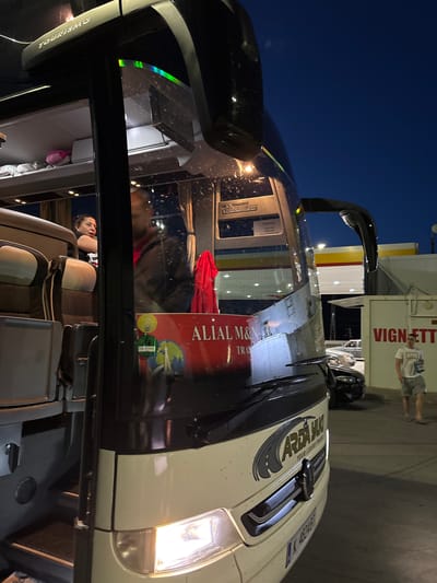 The image depicts the interior of a bus at night, with a person reclining in a seat. A sticker with the words "ALIAL M&M Travel" is visible on the windshield. Outside the bus, a building with the word "VIGNETT" is present, along with a person standing near the building. Based on the sticker and the surrounding environment, this could potentially be located somewhere in Kalotina, Bulgaria.