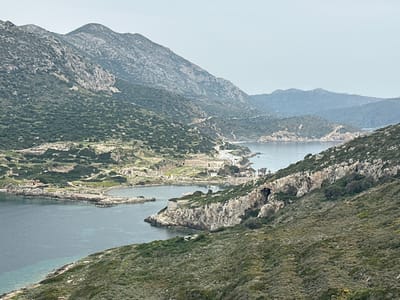 This panoramic image presents a sweeping view of a rugged, mountainous coastal landscape, dominated by the serene waters of a bay or inlet and the extensive ruins of an ancient city. Rolling hills, covered in sparse, scrubby green vegetation and dotted with grey rocky outcrops, rise on either side of the calm, blue-green sea, which appears clearer near the shores. Further mountain ranges recede into the distance under a pale, hazy sky, suggesting an overcast day or atmospheric diffusion.
At the center of the composition, a prominent peninsula juts into the water, hosting the substantial remains of an ancient settlement. Numerous stone foundations, walls, and structures are clearly visible, with what appears to be a large, possibly circular or semi-circular, ancient public building like a theatre or odeon. The layout of the ruins indicates two distinct harbor areas, strategically located for ancient maritime activities. Sections of ancient stonework, including what looks like a fortified wall, can be seen along the rocky coastlines. A winding road is also discernible in the mountainous terrain, suggesting accessibility to the area.
The time of day is daylight, but the diffused light from the hazy sky prevents strong shadows, contributing to a muted color palette. No people or active events are depicted, lending a sense of timelessness and historical depth to the scene. Given the context of Datça, Turkey, the site is almost certainly the ancient city of Knidos, renowned for its two harbors and archaeological significance. The image conveys a sense of peaceful grandeur and the enduring legacy of an ancient civilization amidst natural beauty. No visible text is present in the image.
