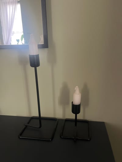 This is a medium shot, eye-level, indoor photograph taken of two black metal candle holders with white candles on a dark surface, likely a table. The candle holders are of different heights, with the taller one to the left and the shorter one to the right. The candle on the left is slightly taller than the one on the right. In the background, a wall painted a light green color can be seen. To the left, a portion of a dark-framed mirror is visible, reflecting a window with white curtains. The overall lighting is soft, creating subtle shadows of the candle holders on the wall. There is no indication of the city of Староживотинное, Russia, within the image itself.