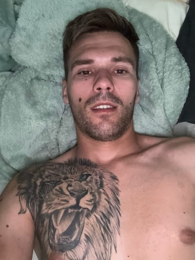 A close-up, overhead shot shows a man lying on a soft, fuzzy, light teal blanket in what appears to be a bedroom. He is looking directly at the camera. His head is slightly tilted to his left, and his hair is short and brown. He has a light beard and stubble. His eyes are open and focused on the camera. A mole is visible on his right cheek, just below his eye. His mouth is slightly open, revealing his teeth. He has a large, detailed tattoo of a roaring lion on his chest and left shoulder. The lion tattoo is rendered in black and grey, with sharp teeth, a fierce expression, and blue eyes. The man's chest and upper torso are visible, with no shirt on. The background consists of the teal blanket and a glimpse of a white pillow and dark fabric. There are no discernible landmarks or clues to indicate a specific location in Sesto Calende, Italy.