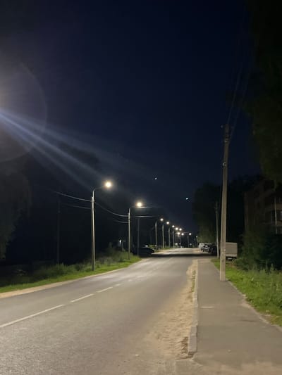 The image depicts a dimly lit street scene at night in Nikolskoye, Russia. A paved road with a white center line stretches into the distance, flanked by a sidewalk on the right and grassy verges on both sides. Several streetlights illuminate the scene, casting pools of light on the road and sidewalk. Trees and vegetation line the sides of the road, and a multi-story building is visible on the right, partially obscured by trees. A few vehicles, including a truck, are parked along the side of the road. The sky is dark, indicating nighttime. The overall impression is of a quiet, ordinary street at night, with artificial lighting providing visibility.
