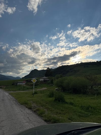 The image depicts a rural landscape under a cloudy sky, with the sun partially obscured by clouds and a hill. A dirt road occupies the left side of the frame, leading towards a small cluster of buildings. Among these buildings, a house with a white facade and a red-roofed structure are visible. A yellow diamond-shaped road sign with a white border stands prominently near the road intersection. The surrounding environment is characterized by green, undulating hills covered in grass and trees. The overall scene suggests a serene, possibly mountainous, rural setting in the region of Verkhnyaya Mara, Russia. No people are visible in the image.