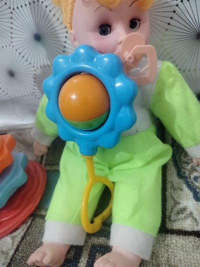 The image shows a close-up of a baby doll sitting on a patterned rug. The doll has blonde hair, blue eyes, and is wearing a light green outfit with white trim on the sleeves and pant legs. It has a pink pacifier in its mouth and is holding a blue and yellow rattle with an orange ball inside. To the left of the doll, a stack of colorful plastic rings, part of a baby toy, is visible. The background consists of a white patterned surface. The image is a still photograph and provides no information about the location in Cholpon, Kyrgyzstan.