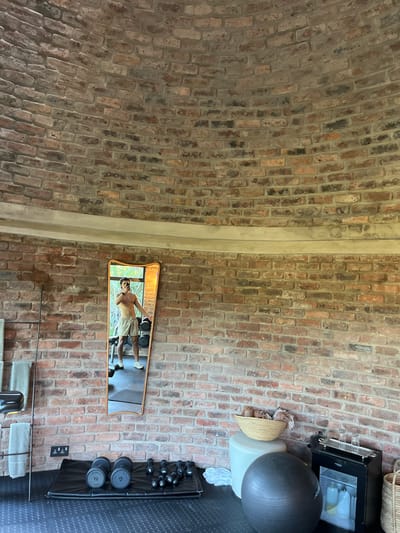 A high-angle shot of a fitness space in Livingstone, Zambia, reveals a blend of rustic charm and modern functionality. The walls and ceiling feature exposed brickwork, creating a warm and textured environment. A vertical concrete pillar divides the composition.

To the right, a small mirror reflects a man, shirtless and wearing shorts, seemingly examining his physique. Below the mirror, a towel rack is placed. Near the top right, a small refrigerator sits atop a basket and next to a gray exercise ball on top of a white canister. 

Weightlifting equipment lines the right edge of the room, a rack with black dumbbells and kettlebells, and a black rubber floor. The lighting is bright, casting soft shadows within the room.