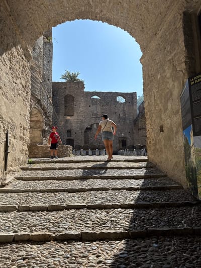 The image captures a view looking through a stone archway towards a cobbled stairway within the medieval village of Dolceacqua, Italy. Two individuals are visible on the stairs. A young boy wearing a red shirt, black shorts, and a red cap stands on a lower step. Further up the stairs, a woman with a light-colored t-shirt and denim shorts walks away from the camera. Behind them, the ruins of a stone structure are visible, with a clear blue sky overhead. A vertical sign with text and an image is positioned on the right side of the frame. The stonework of the archway and the surrounding buildings suggests a historical setting.