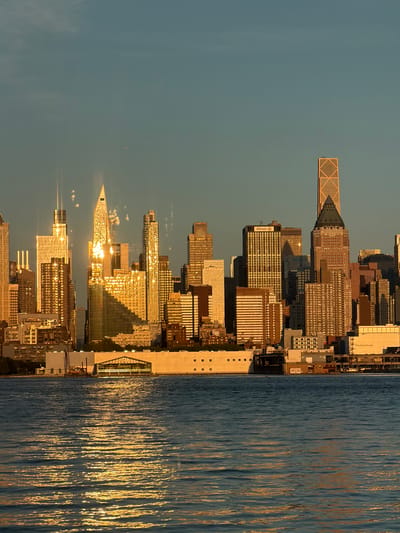 A panoramic view of the New York City skyline is captured from Weehawken, New Jersey, at golden hour. The sun's warm, low light glints off the glass facades of numerous skyscrapers, creating a dazzling spectacle. In the foreground, the calm waters of the Hudson River reflect the vibrant colors of the sunset and the city lights. A large, modern building with a glass-fronted interior and a peaked roof stands prominently along the waterfront. Among the towering structures, an airplane can be seen flying in the distance, a small speck against the vast expanse of the sky. The image depicts a serene and majestic urban landscape at the close of day.