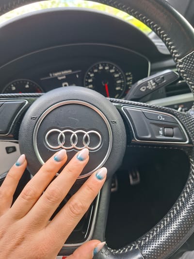 The image displays the interior of an Audi vehicle, specifically the steering wheel and a human hand, located in Rochester, United Kingdom. The black, leather-wrapped steering wheel prominently features the Audi logo—four chrome interlocking rings—in its center. The right spoke of the steering wheel contains integrated controls, including a button with a headset icon and a rotary dial for volume adjustment, indicated by a speaker symbol with plus, minus, and mute lines. To the right of the steering column, a stalk for wiper controls is visible, marked "AUTO." The instrument cluster behind the steering wheel shows a partially visible speedometer on the right with a red needle and a tachometer/fuel gauge on the left. A central digital display shows text, including Arabic script that translates to "speed."
A light-skinned human hand, with manicured nails, rests on the lower left portion of the steering wheel hub. The nails feature an ombre design transitioning from a translucent or pale nude base to a sparkly blue/teal tip. The car's interior is illuminated by natural light, and the blurred background visible through the windshield shows green foliage and a light blue sky, indicating an outdoor daytime setting. Two foot pedals are faintly discernible in the dark footwell.
