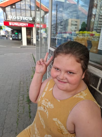A young girl, with brown hair and rosy cheeks, smiles and makes a peace sign with her fingers. She is wearing a yellow dress with a tropical leaf pattern. The background is a bit blurry, showing reflections in a window of a building with storefronts. One prominent sign visible in the reflection reads "SUNLIGHT" in red letters. Another sign is partially visible and says "ROSTIC'S". The ground is paved with grey rectangular stones. The scene appears to be outdoors during the daytime, with a slightly overcast or diffused light. The girl's expression is joyful and playful. The location is identified as Bel'kovo, Russia.