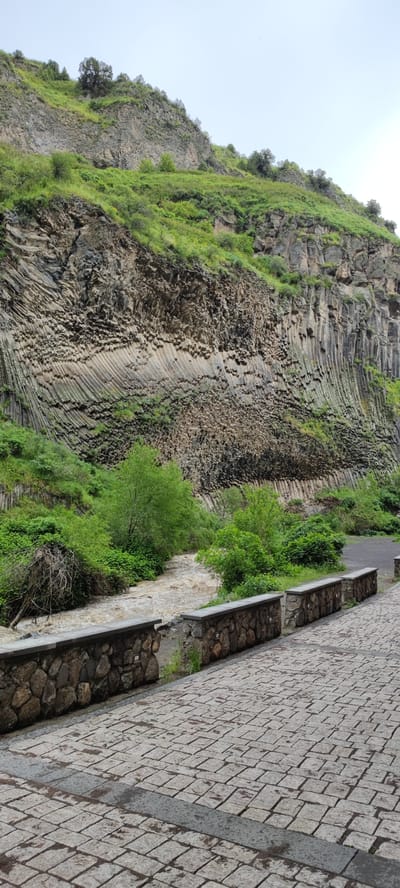 The image depicts a view of the Symphony of Stones, a basalt column formation, within the city of Garni, Armenia. The scene features a river with muddy-colored water flowing alongside a stone-paved walkway. A low stone wall separates the walkway from the river. Green vegetation is visible along the riverbank and on the hillside above the rock formation. The basalt columns display a honeycomb-like pattern in some sections, and vertical columns in others. The sky appears overcast.