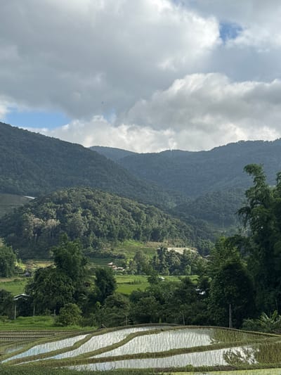 The image depicts a scenic landscape with terraced rice paddies in the foreground, reflecting the sky and surrounding greenery. Beyond the paddies, rolling hills covered in lush vegetation and trees dominate the midground, with several small buildings nestled amongst them. The background features a majestic mountain range shrouded in a light mist, with a cloudy sky overhead. The scene captures the natural beauty of a rural area, possibly in Ban Ang Ka Noi, Thailand, emphasizing the agricultural terraces and the mountainous terrain. There are no people visible in the image.