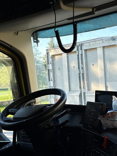The image shows the interior of a vehicle, specifically the driver's side. A black steering wheel is prominently visible. The dashboard and windshield are also in view, with a coiled cable hanging near the windshield. Outside the vehicle, a large metal structure, possibly a trailer or building, is visible. It's difficult to determine the exact location within Umarevtsi, Bulgaria, based solely on this image. The environment appears to be daytime, but without additional information, further details about people, subjects, objects, actions, or interactions are not determinable.