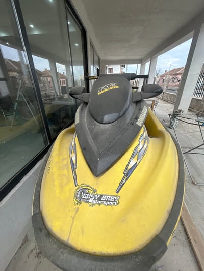 The scene depicts a yellow Sea-Doo RX DI 951 personal watercraft parked on a concrete porch or patio in Dolni Tsibar, Bulgaria. The watercraft, branded "BOMBARDIER" on its side and "RX DI SEA-DOO 951" on its handlebars, displays a "GUSY BABY Motorsport" sticker on its front hull and features black, blue, and white lightning-bolt style decals. It appears dusty and exhibits minor scuff marks. To the left, a large glass wall reflects an outdoor view, revealing traditional houses with red tile roofs, a white A-frame ladder, and a cloudy sky, consistent with a residential area in Dolni Tsibar. To the right, the patio's concrete support pillars frame a background that includes a wrought-iron fence, additional residential structures, and various metal construction materials. A partial view of a white license plate-like object with "01 AE 59" text is visible on the right side of the frame. The lighting is natural and diffused under an overcast sky. No individuals are present.