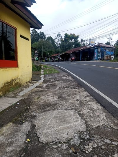 A street scene shows a yellow building with a red-framed window on the left. A dilapidated concrete sidewalk leads to an asphalt road with a white line. Several motorbikes are parked or moving along the road near a worn building with a rusted roof and a sign. Trees and utility poles are visible in the background under a cloudy sky. The sidewalk is cracked and uneven.