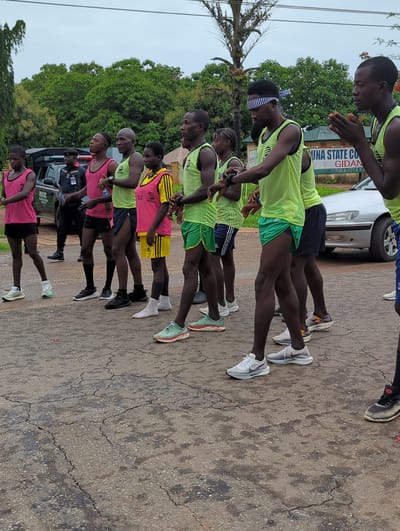 A line of nine black men stand shoulder to shoulder on a cracked asphalt road in Gidan Waya, Nigeria. Most of the men wear running gear consisting of shorts, t-shirts, and running shoes. The runners wear either bright green or bright pink athletic vests, with some wearing yellow vests. Some of the runners also wear headbands. Behind the runners are two cars: one white and one black. A palm tree is visible on the left of the shot. Green trees stand in the background. In the distance, a sign reads "Kaduna State College of Education Gidan". The men are looking down.
