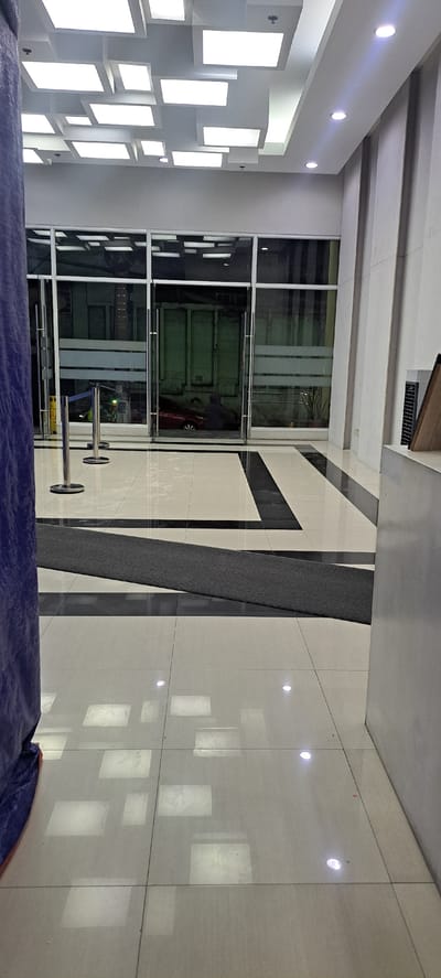 The image shows an interior scene, likely a modern building lobby or hallway, in Mandaluyong, Philippines. The perspective is from a high angle, looking downwards. The scene is dominated by light-colored tiled flooring with a dark accent stripe running diagonally across it. On the left side, a wall is adorned with numerous illuminated square and rectangular panels, creating a striking geometric light display.

Further into the scene, there's a glass-enclosed area, possibly an elevator or a room, with a large green door visible inside. Reflections on the glass show the interior of the space and the surrounding elements. A person in dark clothing and a red object, likely a vehicle or cart, are partially visible within the glass enclosure. In front of the glass enclosure, there are two metal stanchions with ropes, often used for crowd control or marking an entrance.

The ceiling features exposed beams and recessed lighting. The overall impression is one of contemporary architecture with a focus on lighting and reflective surfaces. The time of day is unclear, but the bright interior lighting suggests it's functional and well-lit regardless of external conditions. No discernible text is visible in the image. The scene appears calm and unoccupied, aside from the partially visible person within the enclosed space.