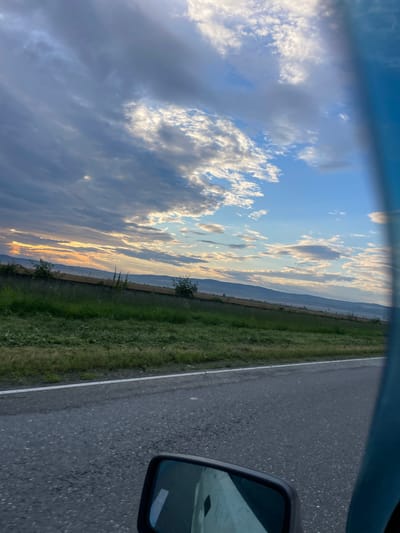 The image depicts a cloudy sky at sunset over a rural landscape. The sun's rays are breaking through the clouds, casting a warm glow on the horizon. In the foreground, a road runs parallel to a grassy field, with a low fence separating the two. The scene appears to be captured from a moving vehicle, as evidenced by the slightly blurred foreground and the reflection in the side mirror. The location is identified as Brut, Russia. The environment is natural and undeveloped, with rolling hills in the distance. There are no people or animals visible in the image. The overall mood is serene and peaceful, with the sunset creating a sense of calm and tranquility.