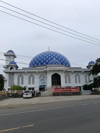The image shows the Masjid Islamic Centre At-Taqarruf in Paya Tringgadeng, Indonesia. The mosque features a large blue dome with a diamond pattern and smaller blue domes on the minarets. The exterior of the mosque is white with decorative latticework on the windows. A banner in front of the mosque reads "MASJID ISLAMIC CENTRE AT-TAQARRUF KAB. PIDIE JAYA TRIENGGADENG". A white car is parked in front of the mosque, and two people are standing near it. The scene is captured on an overcast day. The foreground shows a road with a yellow line. Power lines are visible across the top of the image.