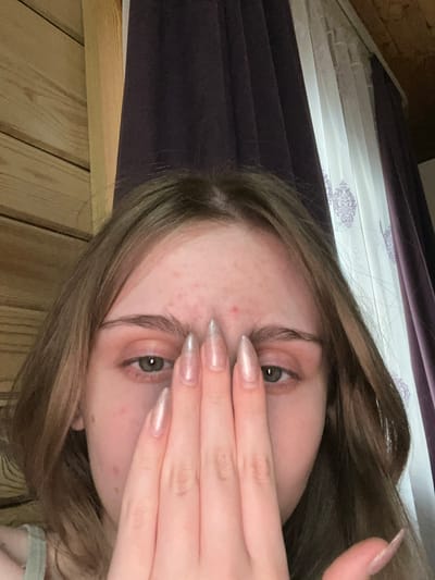 A close-up selfie shows a young person's face partially obscured by their hand. The person has fair skin with some blemishes and light brown hair. They are wearing long, metallic-pink fingernails. The background consists of wooden paneling and draped curtains, suggesting an indoor setting. There is no information in the image to indicate a location in Ashkadar, Russia.