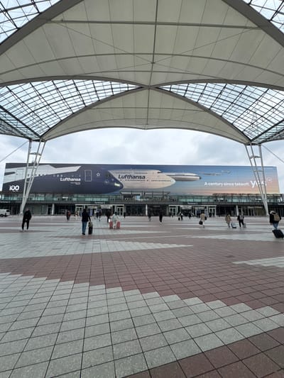 The image shows the entrance of Terminal 2 at Munich Airport (MUC) in Oberding, Germany.
The scene is composed of an exterior view, with a large architectural structure resembling a white canopy supported by metal beams covering a pedestrian area. A prominent advertisement banner depicting a Lufthansa Airbus A350-900 with the Lufthansa logo is visible in the background, partially obscuring the entrance to Terminal 2. The advertisement also displays the text "A century since takeoff" and "Star Alliance."
Several people are walking along the tiled walkway in front of the terminal. The tiled surface is designed with alternating patterns of light gray and reddish-brown tiles. Shop signs, including "Marc O'Polo" are visible behind the advertisement.