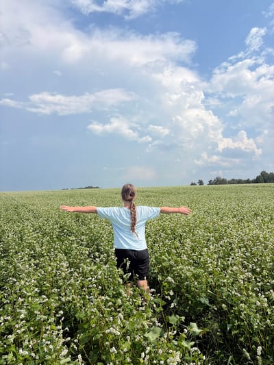 A young person with a braid stands in a field of white flowers, facing away from the camera with their arms outstretched. The field is vast and extends to the horizon. Above, a partly cloudy sky with white cumulus clouds and patches of blue. The location is the village of Nalobikha, Russia.
