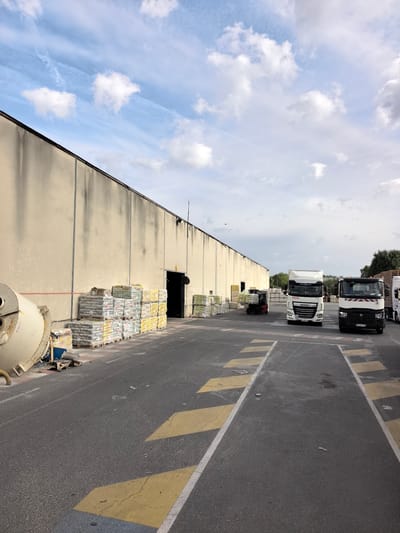 The image depicts a loading dock area outside a large, beige industrial building in Bonneuil-sur-Marne, France. The building has a dark roofline and a row of exterior lights. In front of the building, several pallets of bagged materials, likely cement or similar construction supplies, are stacked and wrapped in plastic. To the left, a large, yellow industrial mixer is positioned.

In the foreground and mid-ground, the asphalt surface is marked with yellow and white directional lines and crosswalk markings. Several trucks are present in the area. Two white semi-trailer trucks are parked, one with "DAF" branding and the other with a red and white striped marking on its cab. A red forklift is also visible near some stacked pallets. The sky is partly cloudy with a mix of blue sky and white clouds. Some trees are visible in the background to the right.