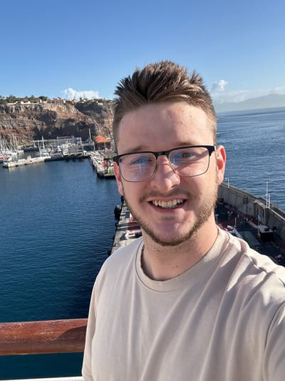 A male subject with light skin, short brown hair styled upwards, a light beard, and dark-rimmed glasses smiles directly at the camera. He wears a plain light beige t-shirt. The subject is positioned in the foreground, with a reddish-brown wooden railing visible in the lower left. The background reveals a harbor and marina situated in San Sebastián De La Gomera, Spain. A deep blue body of water occupies the middle ground. To the left, numerous white recreational boats are docked within a marina. A concrete pier extends from the left towards the right, separating the inner harbor from the open sea. Behind the marina, rugged, reddish-brown cliffs rise steeply, with scattered buildings and vegetation visible on their upper sections. In the far distance to the right, across the water, a distant landmass is visible. The sky is clear blue with a few small white clouds. The scene is brightly illuminated by natural daylight.