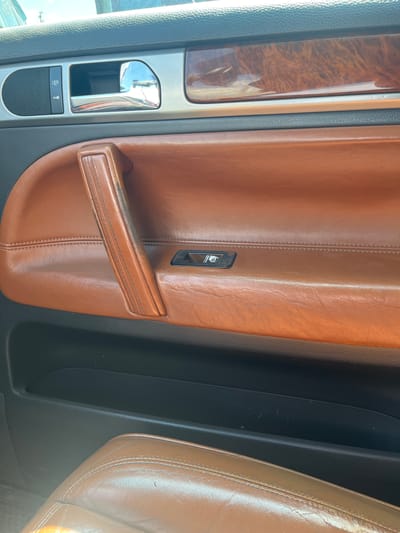 This image shows the interior of a car, likely a luxury model, with a focus on the door panel. The dominant colors are warm browns from the leather upholstery and wood trim, contrasting with the cool metallic finishes of the door handle and controls.
The subjects are the car's interior components. We see a brown leather door panel with prominent stitching, a pull-handle also in brown leather, and a smaller pull tab above it. To the left, there is a section with a wood-grain trim insert, a silver metallic surround, a chrome door handle, and a control panel with buttons and a speaker grille. A single window control button is visible on the brown leather portion of the door. To the right, the edge of a car seat, also upholstered in brown leather, is visible.
There is no activity or event taking place; the image is a still shot of the car's interior.
Notable details include the high-quality materials like leather and wood, suggesting a premium vehicle. The lighting appears to be natural, possibly daylight, casting soft reflections on the metallic surfaces. The location cues are minimal, but the context provided places this car in Aqköl, Kazakhstan. There are no visible emotions conveyed as the image focuses on objects.
There is no visible text in the image, other than the icons on the control panel which are too small to decipher clearly.