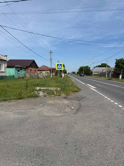 This is a medium shot of a street scene in Nogir, Russia. The road runs from the foreground to the background, curving to the left. On the right side of the road, there are houses and trees. On the left side of the road, there is a grassy area with some debris, houses and a pedestrian crossing sign. The sky is blue with some clouds.

In the foreground, the road is asphalt with white markings. There is a manhole cover on the left side of the road.

In the middle ground, on the left side of the road, there is a grassy area with some weeds and a pedestrian crossing sign. Behind the grass, there are houses with brick and metal roofs, and a green fence.

In the background, the road continues and curves to the left. There are houses and trees on both sides of the road. A dark-colored car is driving on the road.

The image is well-lit and the colors are natural. The focus is on the road and the houses on the side of the road.