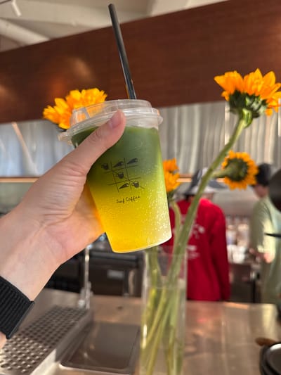 A hand holds a plastic cup of a layered green and yellow beverage with a black straw in front of a vase of sunflowers. The cup has "Surf Coffee" printed on the side. The background appears to be a cafe interior, including metal surfaces and a red object. The image was taken at a location within the city of Кощаково, Russia.