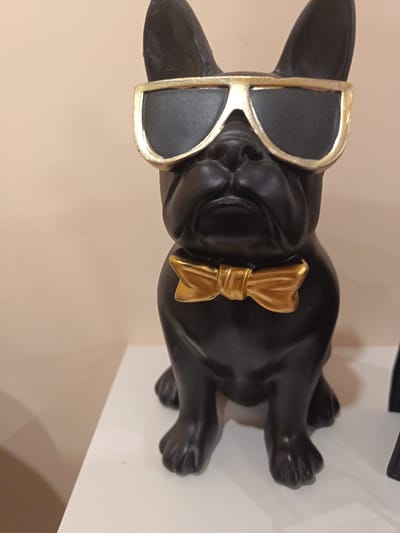A black bulldog statue wearing sunglasses and a gold bow tie sits on a white surface. The background is a plain, light-colored wall. The location is not evident in the image and cannot be determined to be Brezovo, Bulgaria. No people are present.
