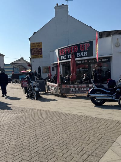 The image depicts a street scene in Walton-on-the-Naze, United Kingdom, featuring a white, two-story commercial building housing "Revved Up The Tea Bar" on the ground floor, identifiable by its black fascia with red lettering and an outdoor seating area. Adjacent to it on the left is another white building with a yellow sign reading "The Naze Fish & Chips," advertising "KEBABS BURGERS CHICKEN PIZZA" and a phone number.
Approximately ten to fifteen individuals are visible. Most are seated in the outdoor area of "Revved Up The Tea Bar," under collapsed red umbrellas, behind a branded barrier displaying "REVVED UP TEA BAR," a Union Jack, and "Jack Daniel's" branding. Many of these individuals appear to be adult males, several wearing dark jackets, possibly leather. A man in a dark jacket and trousers is walking from left to right in the foreground.
Two motorcycles are prominent: a silver/chrome cruiser-style motorcycle with a rider is positioned on the left side of the outdoor seating area, facing right. The rear section of a dark-colored cruiser-style motorcycle with chrome exhaust pipes and a sissy bar is visible on the far right.
The foreground consists of a paved surface, transitioning from a herringbone pattern of grey rectangular blocks to larger square slabs. Two A-frame signs are positioned on the pavement: one black with a "Coca-Cola" logo, and another with "Tea Is Green" text. The scene is illuminated by strong sunlight, casting distinct shadows on the pavement under a clear blue sky.