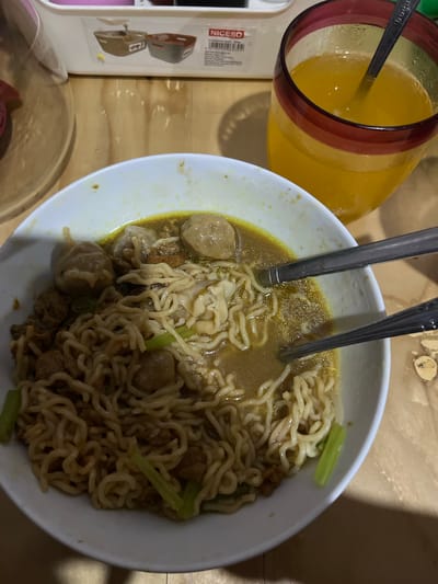A top-down, slightly angled medium shot captures a bowl of instant noodles with meatballs and a glass of orange juice on a wooden surface. The noodles, a mix of wavy and straight strands, are submerged in a yellowish broth. Several round, pale meatballs are scattered throughout the bowl, along with small pieces of what appears to be minced meat and chopped green vegetables, possibly scallions or celery. Two metal spoons rest in the bowl, one with its handle extending out of frame and the other partially submerged. To the right of the bowl, a tall glass filled with a vibrant orange liquid, presumably juice, sits tilted. The glass has a dark red rim and a dark red band around its middle. A spoon is also present in the glass. In the background, a white plastic container with a label that reads "NICESO" is visible, depicting images of small storage containers. The wooden surface has a visible grain and appears to be a table or countertop. The lighting suggests an indoor setting, potentially with natural light contributing to the overall illumination. The scene is devoid of any human presence. The location is Serdang Kulon, Indonesia.