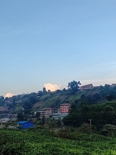 A landscape displays a hillside dotted with buildings of various sizes and colors, including a structure with a red facade. Lush vegetation lines the right side of the frame. Power lines stretch across the hill, and a portion of a blue-roofed building is visible. The sky occupies a significant portion of the view, showcasing a gradient of light blue.