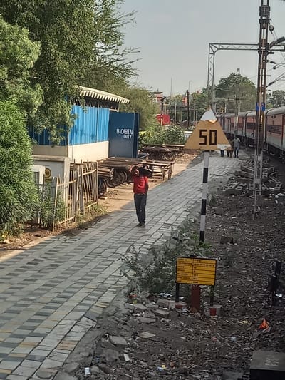 The image depicts a railway scene likely in Bhopal, India. A man in a red shirt and dark pants walks along a paved walkway. To the left, there's a blue structure labeled "B. CABIN DUTY," partially obscured by trees. Railway tracks and a train are visible in the background, along with overhead power lines. A triangular yellow sign with "50" is displayed, next to a sign with a yellow table on it. Debris and gravel litter the ground beside the walkway.