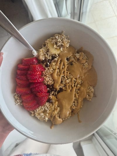 A white ceramic bowl containing a food item is depicted from a top-down perspective, held by an individual's left hand, visible in the lower left quadrant. The bowl's contents include a base of textured oats or granola, topped with a row of sliced red strawberries on the left side, interspersed banana slices, scattered dark chocolate chips, and a generous drizzle of light brown peanut butter covering the right side. A silver spoon is partially inserted into the bowl on the upper left, with a small quantity of white substance, possibly yogurt or cream, on its tip. The background is out of focus, featuring light-colored curtains or fabric on the left and top, and a window or light source with a sheer screen on the right, indicating an indoor environment under natural light. The specific location within the Municipal Unit of Corinth, Greece, cannot be determined from the visual evidence provided.