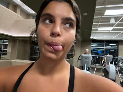 The image depicts a young adult female captured in a close-up, selfie-style photograph, positioned in the foreground of an indoor gymnasium. She has dark hair, brown eyes, and tan skin. Her eyes are directed upwards and to the right, while her tongue is extended from her mouth. She is wearing a black athletic top and small hoop earrings. In the background, typical gym architecture is visible, including light-colored walls and a suspended ceiling with recessed lighting panels. Large windows show an obscured exterior, likely indicating nighttime. Two other individuals are present in the mid-ground, operating exercise equipment. One bald male with glasses is visible on an elliptical or stationary bike, looking forward. A second, partially obscured individual is also on an exercise machine. The scene is illuminated by artificial indoor lighting. The location is Plainsboro Township, United States.